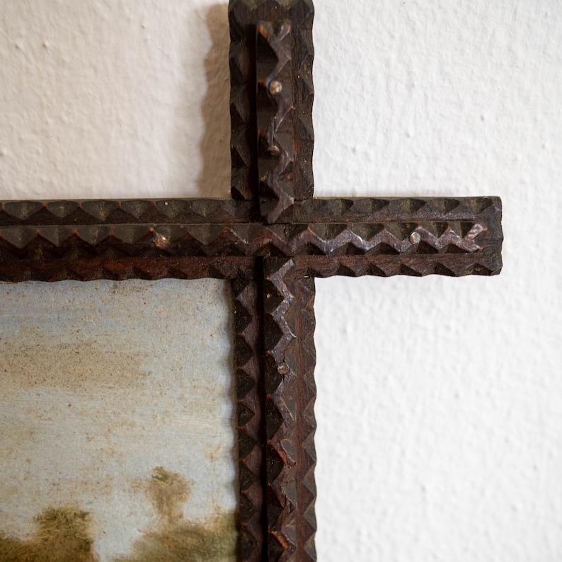 May include: A rustic, dark brown wooden cross-shaped frame with a landscape painting. The frame has a textured, carved design along the edges. The painting features a muted color palette with a landscape scene.
