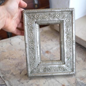 Può includere: Una cornice rettangolare color argento con dettagli floreali e di vite. La cornice ha un bordo rialzato con un motivo ripetuto di fiori e foglie. La cornice è tenuta in mano.