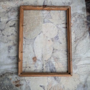 Può includere: Una cornice rettangolare in legno con finitura naturale. La cornice è vuota, rivelando uno sfondo di carta strutturata con impronte di foglie nei toni del bianco, grigio e marrone. La cornice è fissata con piccoli chiodi.