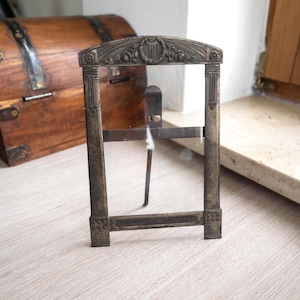 May include: An antique metal picture frame with a clear glass insert. The frame has an arched top with floral and wreath detailing. The frame is rectangular with vertical columns on each side and a horizontal base. The frame is a muted bronze color.