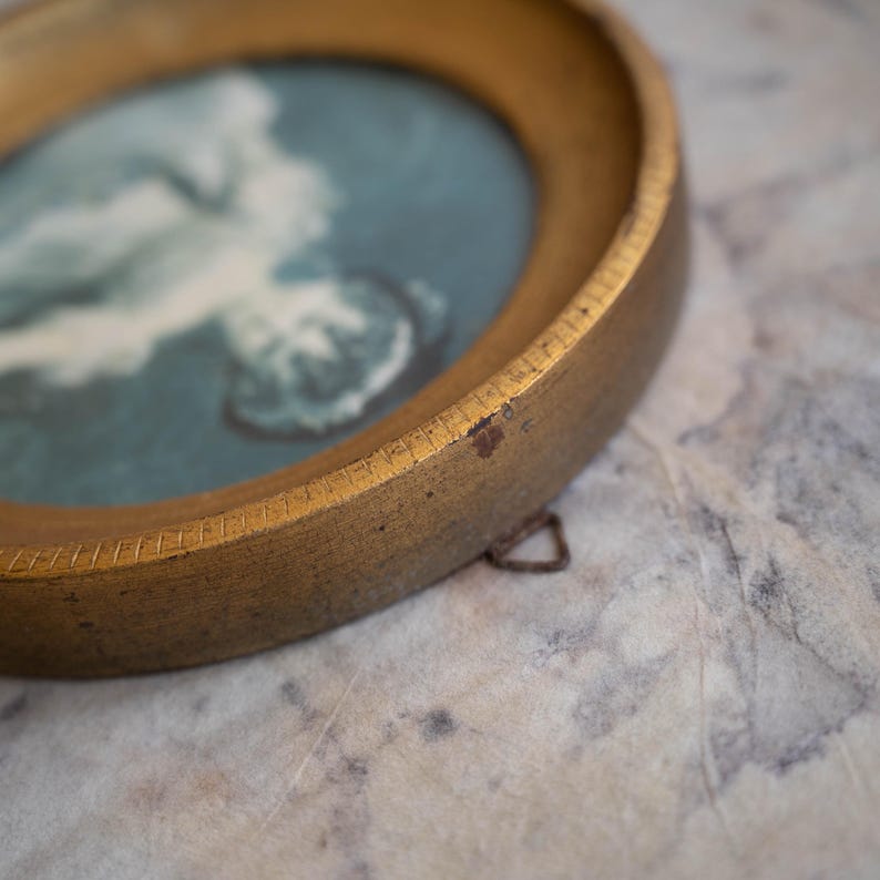 May include: A gold-colored round picture frame with a blue and white image inside. The frame has a small metal hook on the back.