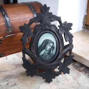 Könnte beinhalten: Ein schwarzer, geschnitzter Holzbilderrahmen mit einem ovalen Porträt einer Frau mit Kopfbedeckung. Der Rahmen hat ein Blattdesign. Das Porträt ist in Graustufen gehalten. Im Hintergrund befindet sich eine Holzkiste.