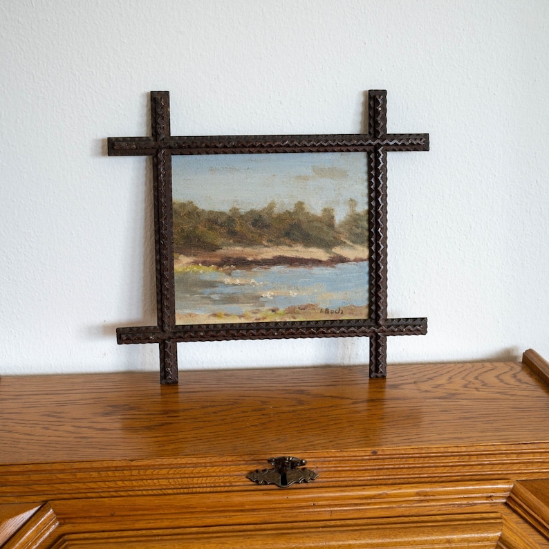 May include: A small landscape painting in a dark brown, intricately carved wooden frame. The painting depicts a coastal scene with a blue sky, water, and green trees. The frame has a cross-like design at each corner.