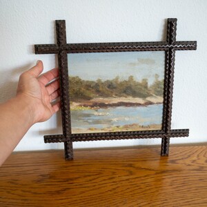 May include: A small landscape painting in a dark brown, intricately carved frame. The artwork depicts a serene waterside scene with a light blue sky, water, and green trees. The frame has a cross-like design at each corner.