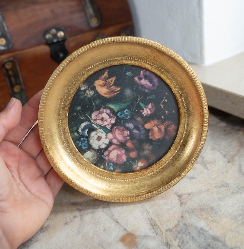 A hand holding the round gold frame and gilded wooden photo frame displaying a dark floral print.