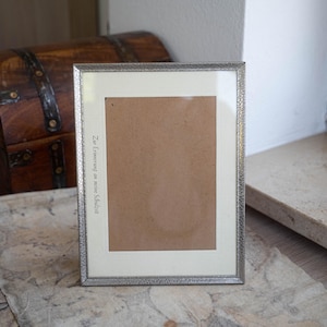 Può includere: Una cornice argentata con un bordo strutturato. La cornice contiene un passe-partout marrone e un bordo color crema. Il testo "Zur Erinnerung an meine Schulzeit" è stampato verticalmente a sinistra. Un baule di legno è visibile sullo sfondo.
