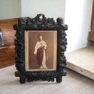 May include: A vintage black framed religious print featuring a sepia-toned image of Jesus Christ. The frame is intricately carved with floral and leaf motifs. The print is displayed on a light-colored surface.