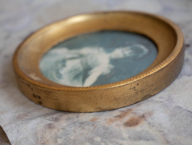 May include: A gold-toned oval picture frame with a faded image of a person inside. The frame has a decorative edge.