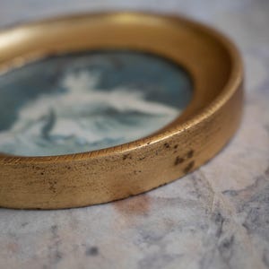 May include: A gold-toned round picture frame with a blue and white image of a person inside. The frame has a decorative edge.