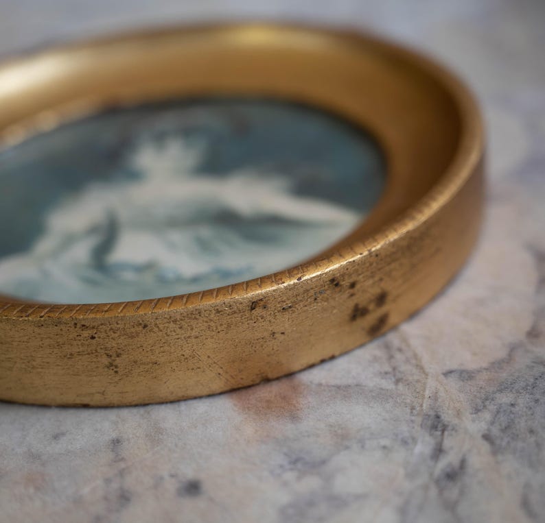 May include: A gold-toned round picture frame with a blue and white image of a person inside. The frame has a decorative edge.