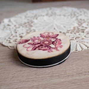 May include: A round, vintage-style compact with a floral design in shades of pink and purple. The compact has a black band around the edge and a white base. The floral design is embroidered on a cream-colored fabric.