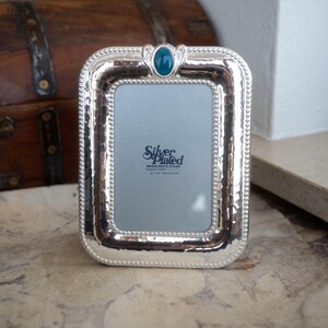 May include: A silver-plated brass photo frame with a hammered texture and a beaded border. The top center features an oval teal cabochon accent. The insert reads Silver Plated Brass Photo Frame 3in x 5in 8cm x 13cm. The frame has a rectangular shape.
