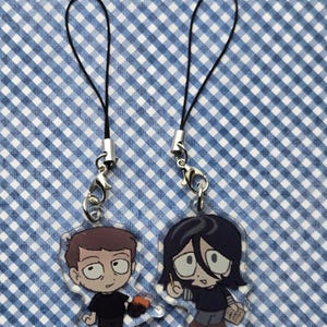 以下が含まれることがあります： 黒い紐のついた透明なアクリルチャームが2つ。チャームには2人の人物の漫画イラストが描かれている。一人は黒いシャツを着て赤い物体を持ち、もう一人は青いシャツを着て鉛筆と紙を持っている。