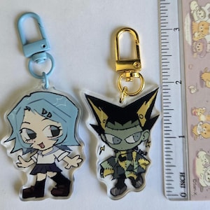May include: Two acrylic keychains featuring cartoon characters. The keychain on the left features a blue-haired girl wearing a white and black school uniform. The keychain on the right features a green-skinned monster with black hair and a yellow and black outfit.