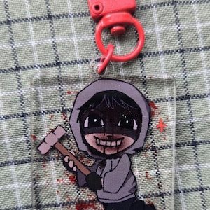 May include: A clear acrylic keychain with a red clasp. The keychain features a cartoon illustration of a person in a gray hoodie holding a hammer, with blood splatter details. The character has a sinister grin.
