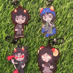 May include: Four acrylic keychains featuring cartoon characters with horns and various outfits. Each keychain has a black metal clasp. The characters have different hair colors, outfits, and expressions. The keychains are displayed on a bed of green grass.