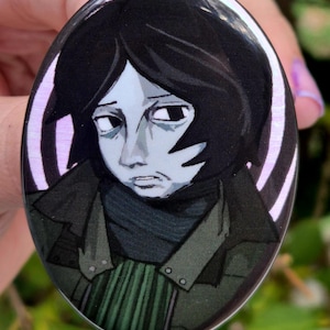 May include: Oval button with a glossy finish, depicting a person with black hair and a melancholic expression. The person is wearing a dark green jacket and scarf. The button has a light purple and white spiral border.