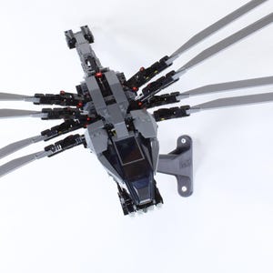 May include: A gray and black model helicopter with long, thin blades extending from the top. The helicopter has a detailed design, including a cockpit and various mechanical components. The blades are a light gray color, and the body is primarily dark gray.