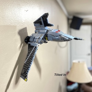 May include: A gray and black Lego Star Wars TIE Interceptor spaceship is mounted on a wall. The spaceship is tilted slightly to the right. The text "Tilted In" is written below the spaceship.