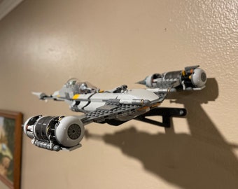 May include: A white and grey Lego Star Wars spaceship with yellow accents is mounted on a wall. The spaceship has two engines at the back and two engines on the bottom.