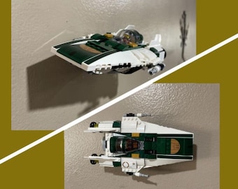 May include: A white and green Lego spaceship with gold accents. The spaceship has a cockpit and two wings. The spaceship is on a white background.