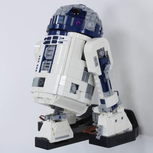 Puede incluir: Modelo R2-D2 blanco, azul y gris construido con ladrillos de plástico entrelazados. El modelo está montado sobre una base negra y presenta elementos mecánicos detallados. La cúpula es gris y azul, y el cuerpo es principalmente blanco.