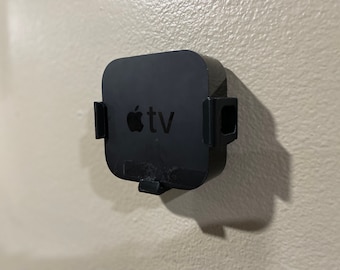 Op de afbeelding: Een zwart Apple TV streamingapparaat gemonteerd aan een muur met een witte achtergrond. Het apparaat heeft een zwart Apple-logo en de tekst "tv" aan de voorkant.