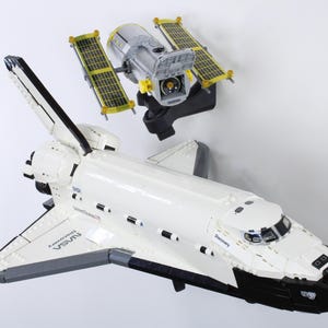 May include: A white and black space shuttle model with the word "Discovery" printed on the side. A satellite model with yellow solar panels is positioned above the shuttle. The models are set against a white background.