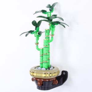 Puede incluir: Una planta de bambú decorativa verde y marrón en una maceta. La planta es de plástico y tiene varios tallos y hojas. La maceta es de color marrón claro con una banda dorada alrededor de la parte superior y se asienta sobre una base negra.