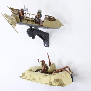 Op de afbeelding: Een beige model van een voertuig met figuren, gemonteerd op een zwarte beugel, boven een beige model van een wezen met bruine tentakels. De modellen worden tegen een witte achtergrond getoond.