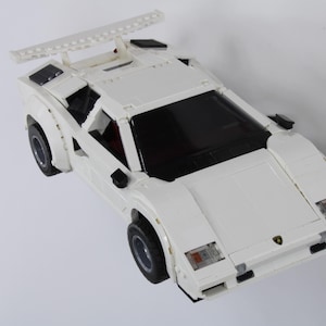 May include: A white toy car with a spoiler and black wheels. The car has a sleek design with a black windshield and headlights. The car has a Lamborghini logo on the front.
