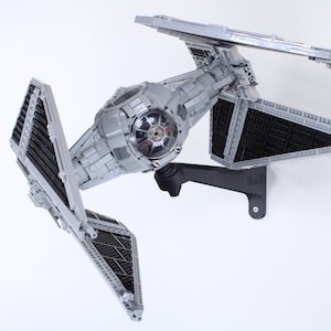 Puede incluir: Un modelo de caza TIE gris con detalles negros está montado en un soporte de pared negro. El modelo está hecho de plástico y tiene un diseño detallado.