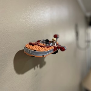 May include: A Lego model of a red and orange spaceship with a minifigure pilot. The spaceship is flying in mid-air against a white wall.