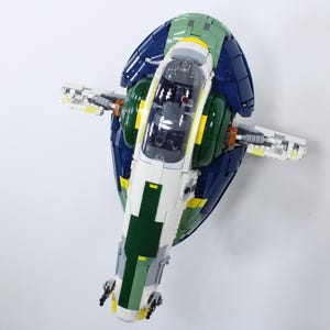 Puede incluir: Maqueta detallada de una nave espacial, con una combinación de colores blanco, verde, azul y amarillo. El modelo tiene alas y una cabina central con una cubierta transparente. El diseño es intrincado, con detalles mecánicos visibles.