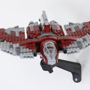 Puede incluir: Una nave espacial de juguete roja, gris y plateada hecha de bloques entrelazados, montada sobre un soporte negro. La nave espacial tiene una cabina y estructuras en forma de alas. El soporte está fijado a una superficie blanca.