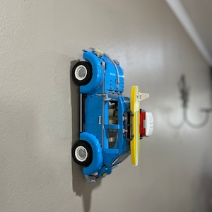 May include: A blue Lego Volkswagen Beetle with a yellow surfboard attached to the roof. The car is mounted on a wall.