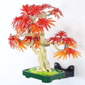 May include: A Lego bonsai tree with vibrant red and orange leaves. The tree has a light tan trunk and sits in a green rectangular pot with a black stand. The leaves are a mix of red and orange.