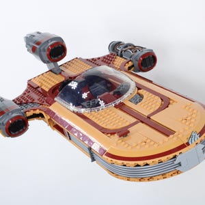 Op de afbeelding: Een beige en bordeauxrood ruimteschipmodel gemaakt van speelgoedblokken. Het model heeft een doorzichtige cockpit, meerdere motoren en een gedetailleerd ontwerp. Het speelgoed is ontworpen om mee te spelen en tentoon te stellen.