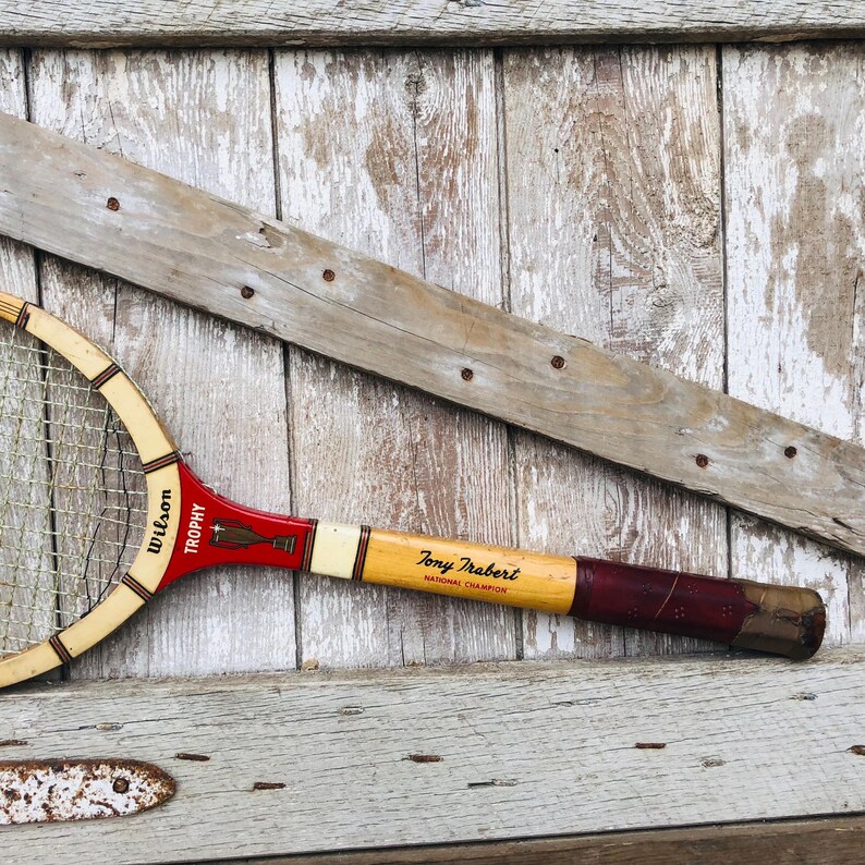 May include: A vintage Wilson Trophy tennis racket with a red, white, and yellow handle. The handle is labeled "Tony Trabert National Champion".