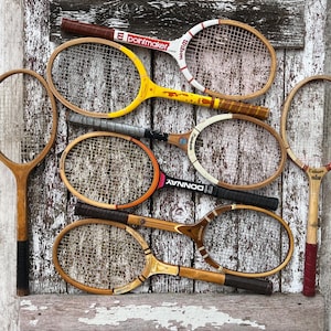 May include: A collection of six vintage wooden tennis rackets with various colored grips, frames, and string patterns. The rackets feature different brand names including Wilson, Pointmaker, and Donnay, displayed with worn wooden textures and metal hardware.