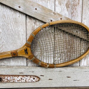May include: A vintage wooden tennis racket with a worn, brown, stringed net. The racket has a dark brown handle and is resting on a weathered wooden surface.