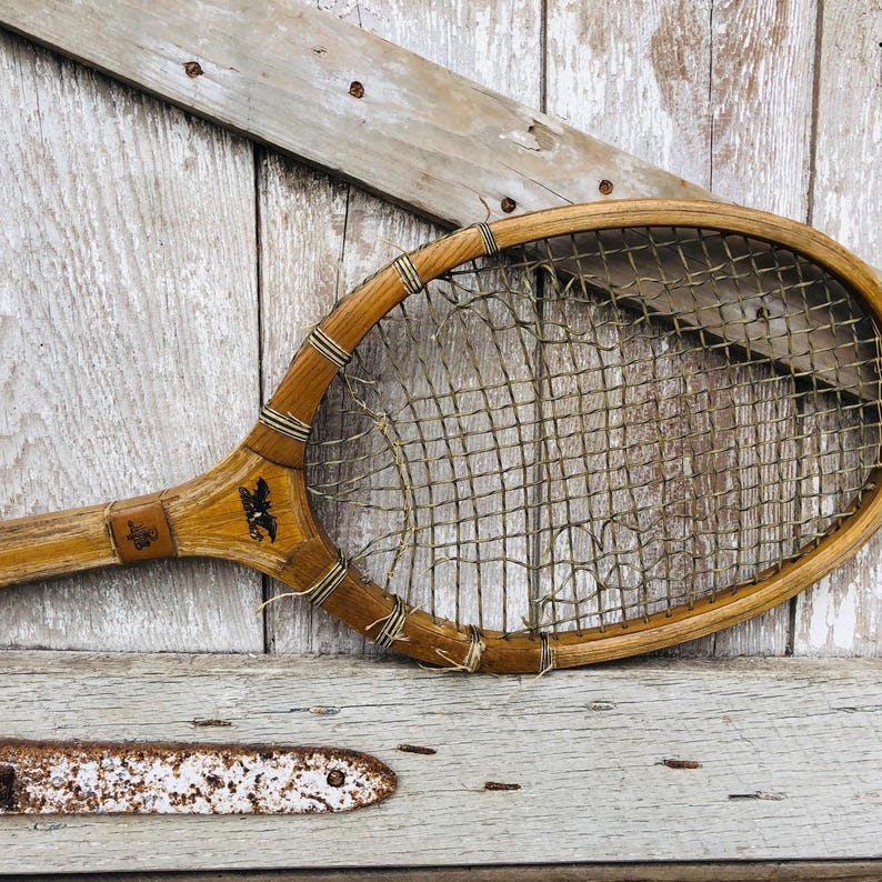 May include: A vintage wooden tennis racket with a worn, brown, stringed net. The racket has a dark brown handle and is resting on a weathered wooden surface.