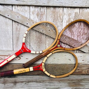 May include: Three vintage wooden tennis rackets with red and white string. The rackets are leaning against a weathered wooden wall. The rackets are labeled "Wright & Ditson" and "Wilson Trophy".