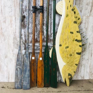 May include: Six wooden oars with different colored paint, hanging on a white wood wall with a yellow and white painted fish hanging below them.