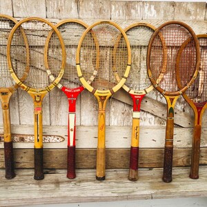 May include: Seven vintage wooden tennis rackets with stringed heads. The rackets are arranged vertically against a wood panel background. The rackets have different colored grips and some have writing on them.