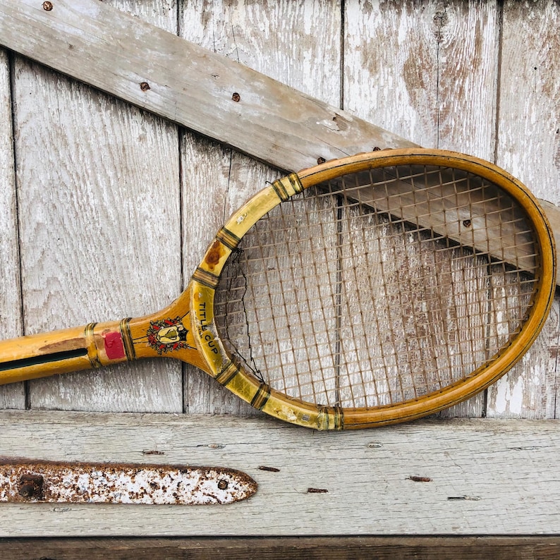 May include: Vintage wooden tennis racket with a yellow and brown finish. The racket has a stringed head and a wooden handle. The handle has a red and gold sticker with the text "Little Cup".
