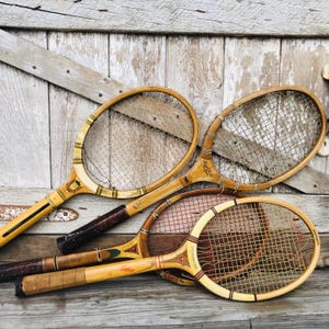 May include: Three vintage wooden tennis rackets with brown handles and yellow and brown stringing. The rackets are arranged on a light wood surface.