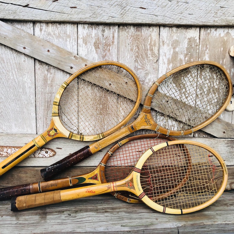 May include: Three vintage wooden tennis rackets with brown handles and yellow and brown stringing. The rackets are arranged on a light wood surface.