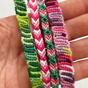 May include: Four woven friendship bracelets featuring intricate knotted patterns. The first is green and lime, the second is pink and white chevron, the third is a green and pink diamond pattern, and the fourth is a multi-colored pink and purple stripe.