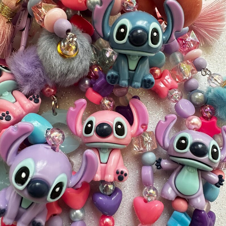 May include: A close-up of colorful beaded accessories featuring Stitch figurines in blue, pink, and purple. The beads are in various shapes and colors, including hearts, spheres, and stars. The accessories also include fluffy pom-poms and other decorative elements.